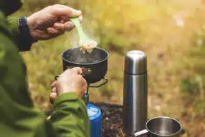 Henkilö laittamassa ruokaa metsässä kaasukeittimellä. Kaasukeittimen vieressä on muki ja termospullo.