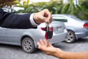 Henkilö antamassa auton avaimet toiselle henkilölle. Taustalla on kaksi autoa.