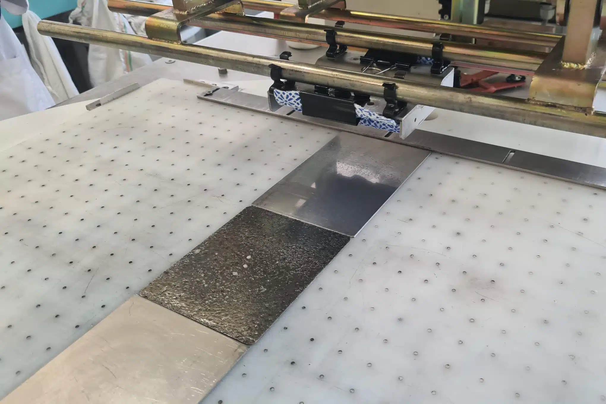 Eri pintamateriaalista valmistettuja suorakulmion muotoisia paloja kiinnitettynä pöytään ja testauslaitteistoa.
